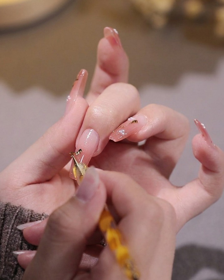 Eine Hand platziert mit einem Dotting-Tool kleine Stücke Blattgold auf den rosa lackierten Nagel.