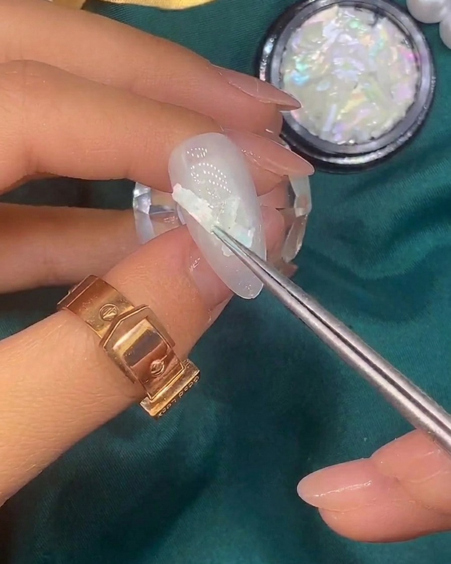 Ein Pinsel trägt winzige Blattgoldflocken auf die Oberfläche eines bereits mit Muschelstücken verzierten Nagels auf.