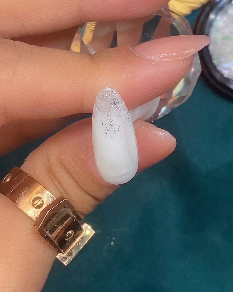 Eine Pinzette platziert ein kleines, unregelmäßiges Stück Silberfolie auf einem Nagel mit silbernem Glitzerverlauf.