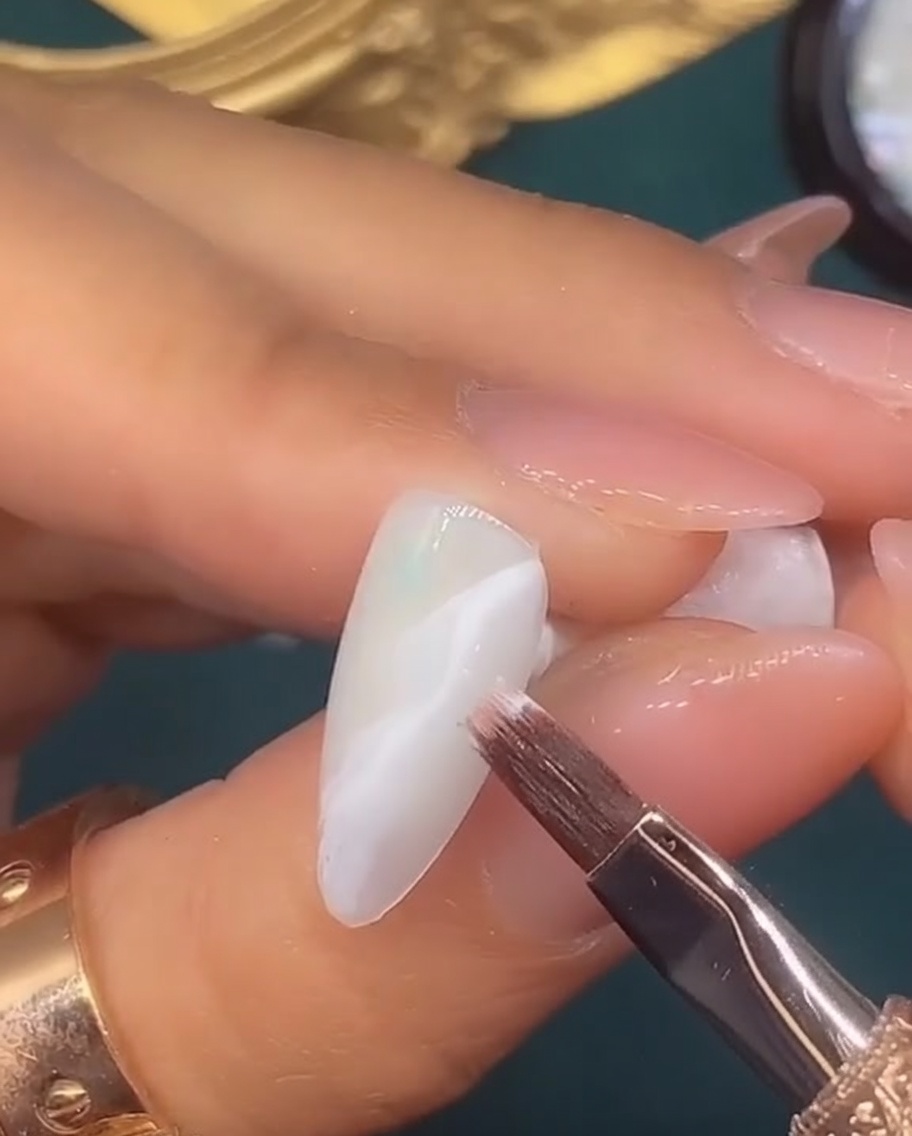 Ein feiner Liner-Pinsel zeichnet ein dünnes, metallisches Wirbelmuster auf einen weiß verblendeten Nagelhintergrund.