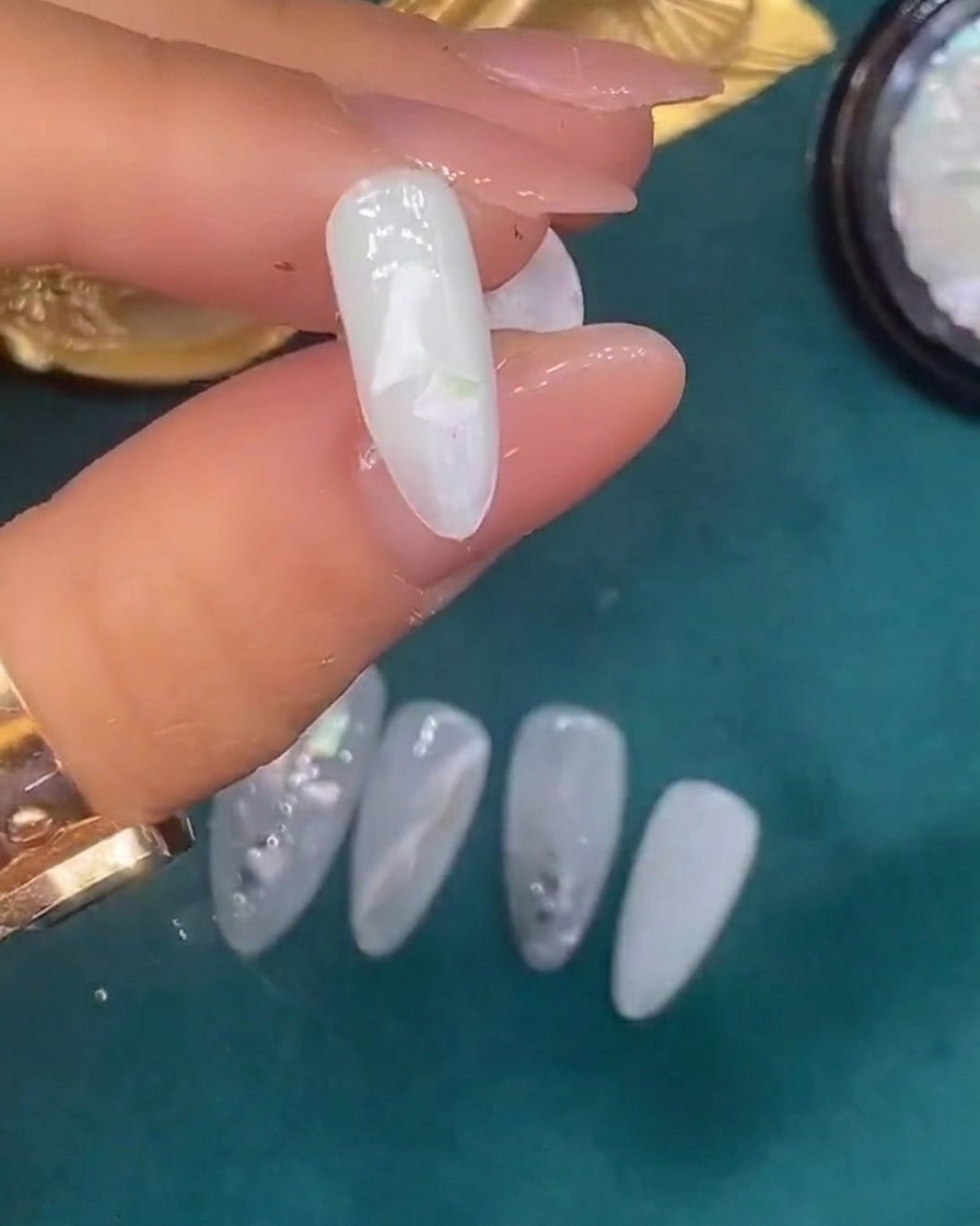 Eine Pinzette platziert dekorative Muschelstücke auf einem mit nassem, durchscheinendem weißem Gel überzogenen Nagel.
