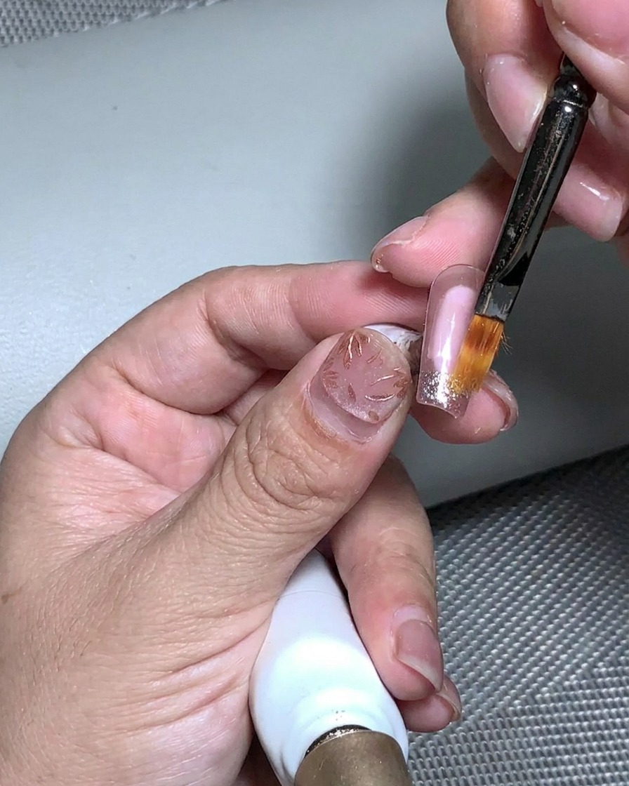 Ein Pinsel, der klares Aufbaugel über die strukturierte Blattgold- und Glitzeroberfläche aufträgt.
