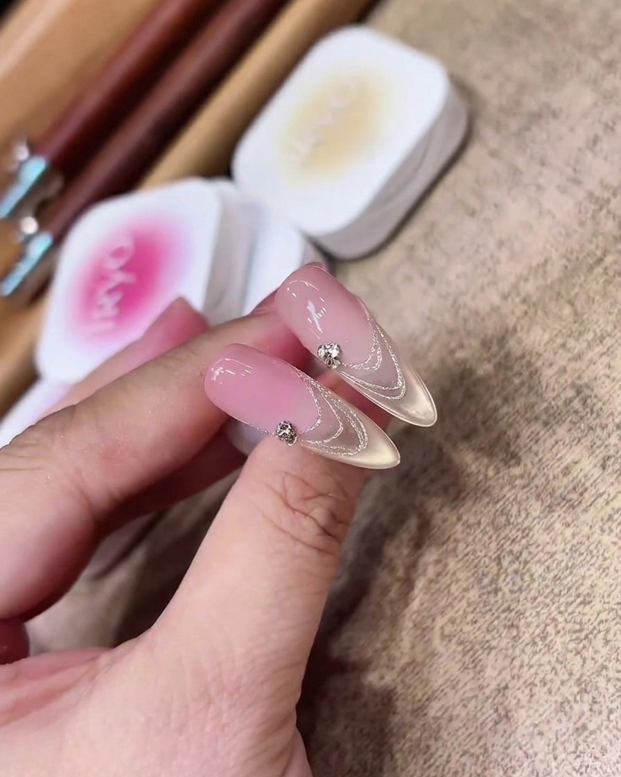 Ein Flachpinsel trägt gleichmäßig ein transparentes, rosafarbenes Nude-Gel auf eine klare, mandelförmige Nagelverlängerung auf.