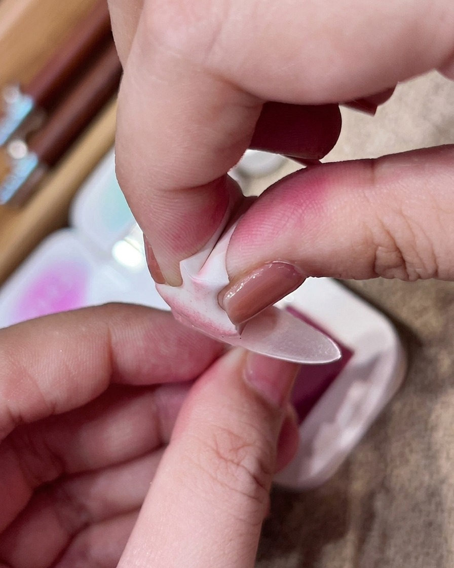 Eine Hand nutzt einen kleinen Make-up-Schwamm, um sanft pinkes Pigment auf die Mitte eines matten Nude-Nagels zu tupfen.