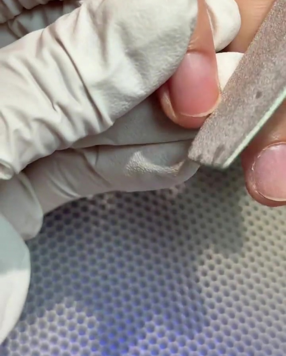 Eine Hand in Handschuhen feilt die freie Kante eines Naturnagels.