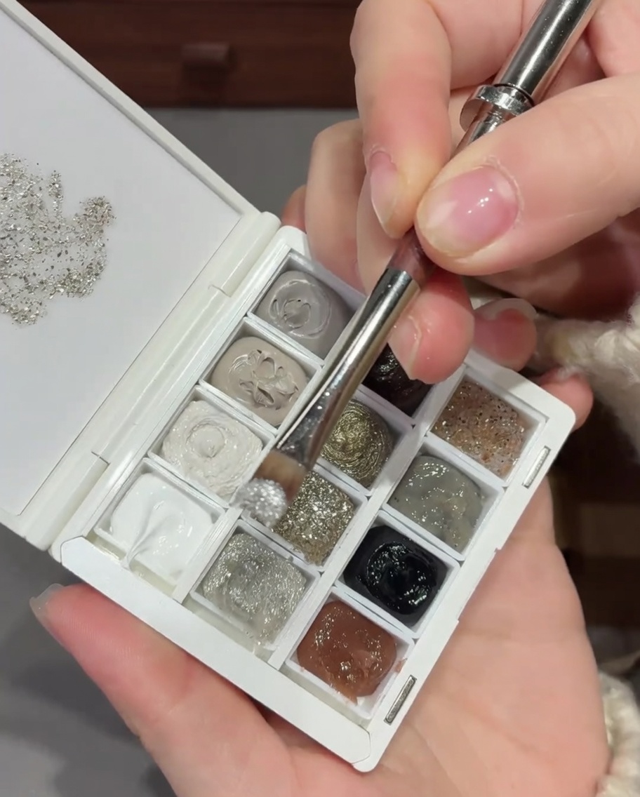 Ein Nagelpinsel trägt eine Mischung aus Silberglitzer und Pailletten an der Basis eines Naturnagels auf.
