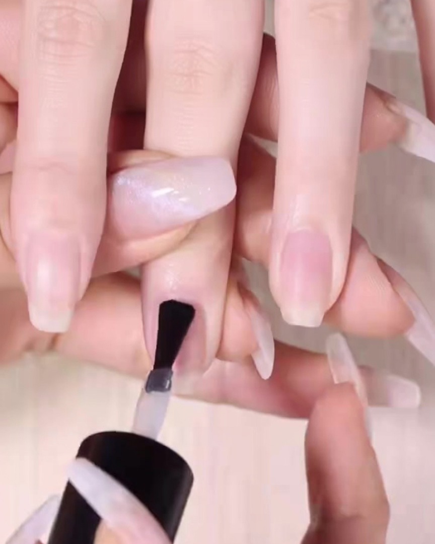 Ein Nagelpinsel trägt eine gleichmäßige Schicht klaren Unterlack auf einen vorbereiteten Naturnagel auf.