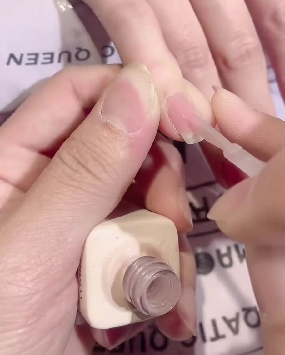 Ein Pinsel trägt eine glatte, dünne Schicht transparenten, nudefarbenen Base-Coat auf einen Fingernagel auf.