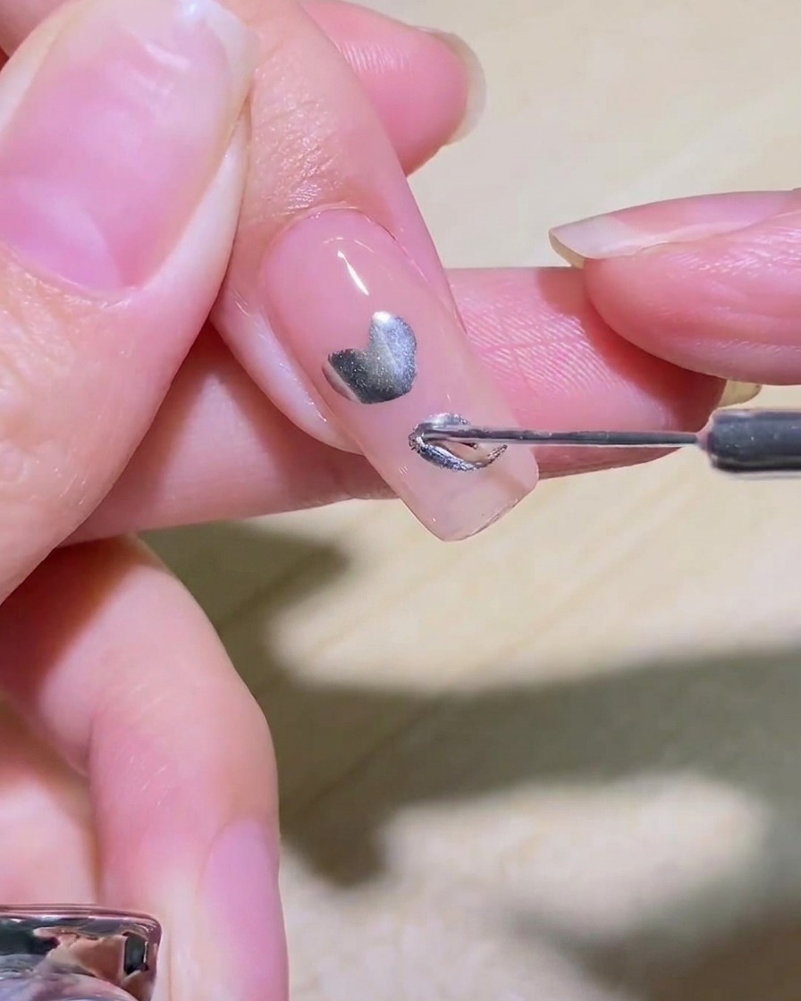 Ein Liner-Pinsel füllt das Innere eines silbernen Herzdesigns auf einem Nagel aus.