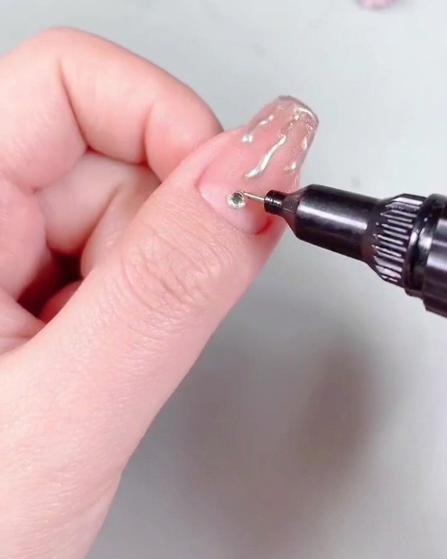Ein Nailart-Pinsel trägt eine Schicht hochglänzenden Stahl-Top Coat über einen Nagel auf, der mit Flammen-Art und einem Strassstein versehen ist.