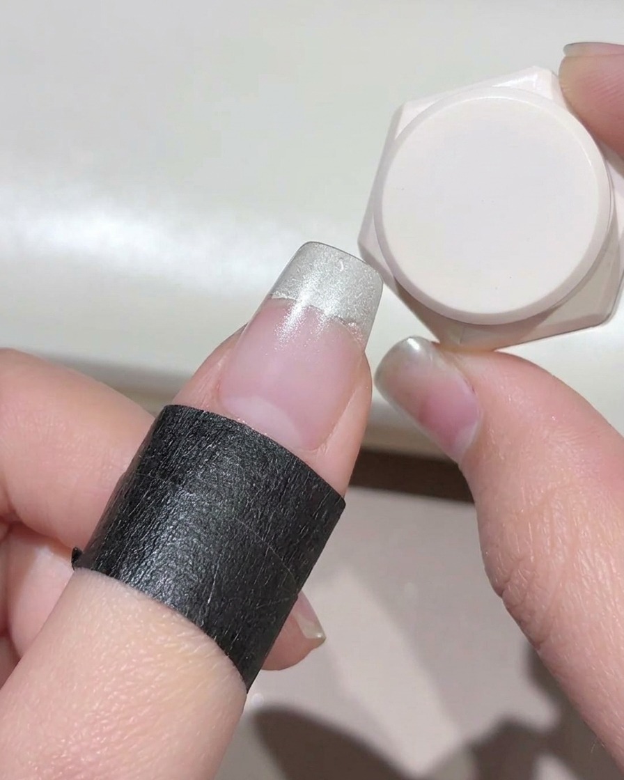 Nahaufnahme eines Fingers mit schwarzem Schutzband um die Haut, wobei milchig-weißes Gel auf die Nagelspitze aufgetragen wird.
