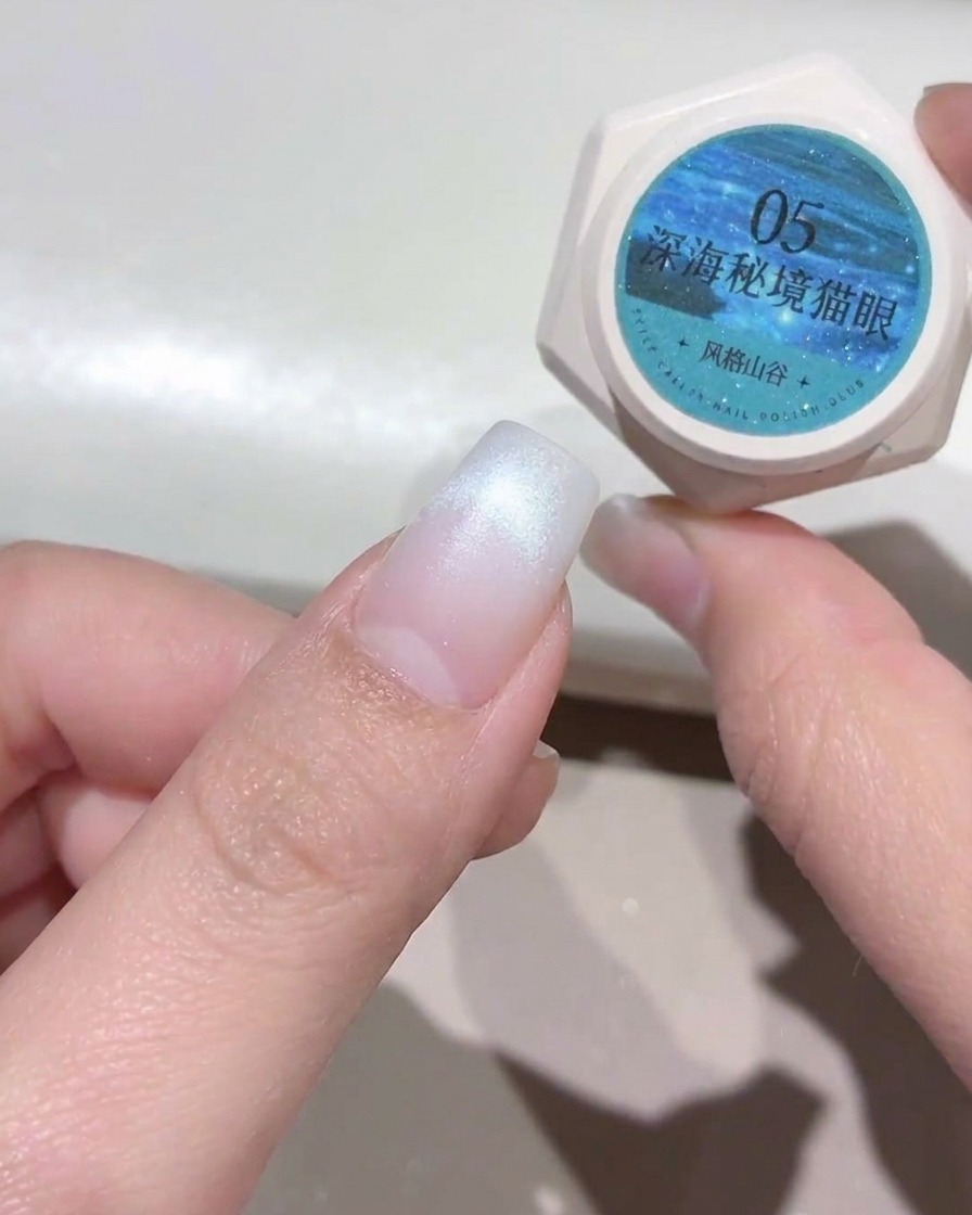 Eine Hand hält einen kleinen Tiegel mit türkis-blauem Cat-Eye-Gel neben einen vorbereiteten Fingernagel mit weißer Spitze.
