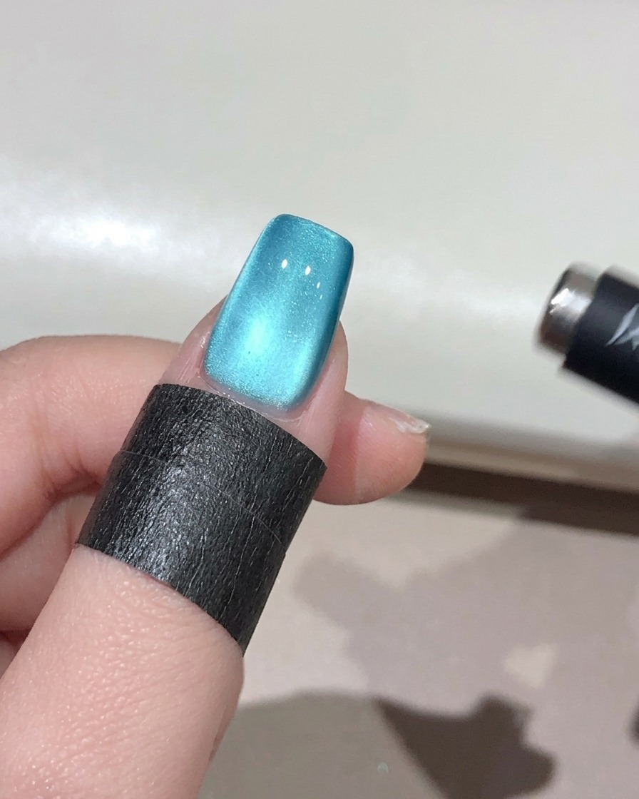 Ein kleiner kugelförmiger Magnet wird in der Nähe des Randes eines türkisen Nagels gehalten und konzentriert den Schimmer zu einem leuchtenden 3D-Perleneffekt.
