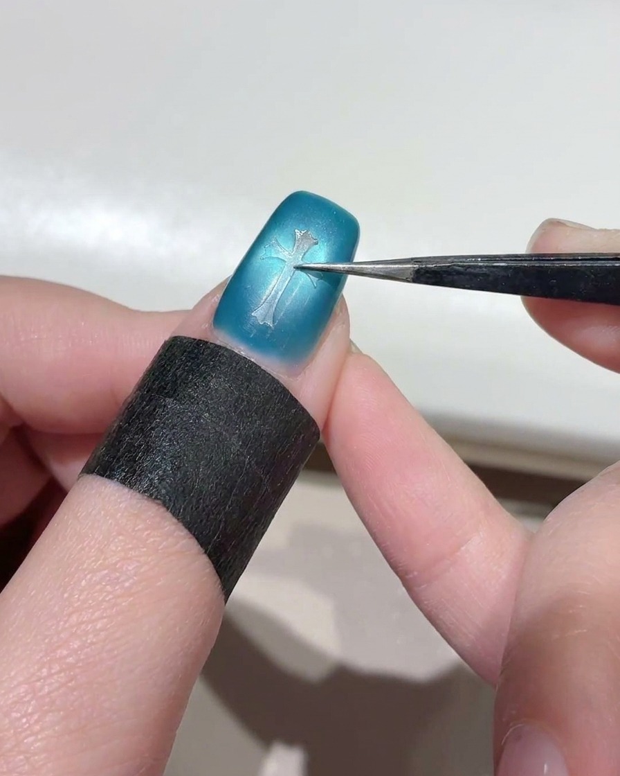 Eine Nahaufnahme zeigt eine Pinzette, die einen klaren kreuzförmigen Schablonenaufkleber auf die Mitte eines schimmernden blauen Cat-Eye-Nagels drückt.