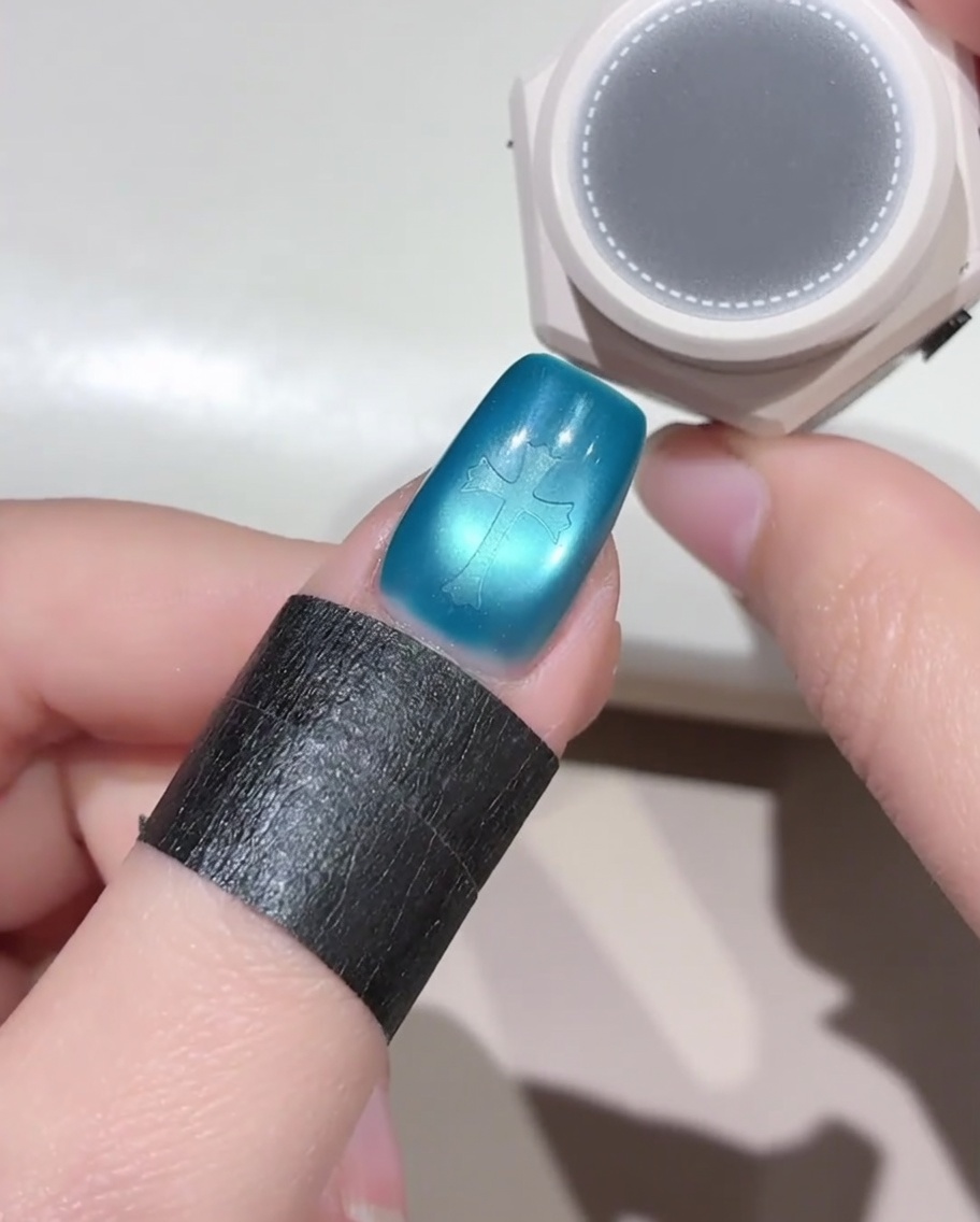 Ein kleines, rundes Schwämmchen schwebt über einem blauen Fingernagel, auf dem sich ein Kreuz-Aufkleber befindet, und bereitet das Auftragen von schwarzem Gellack vor.