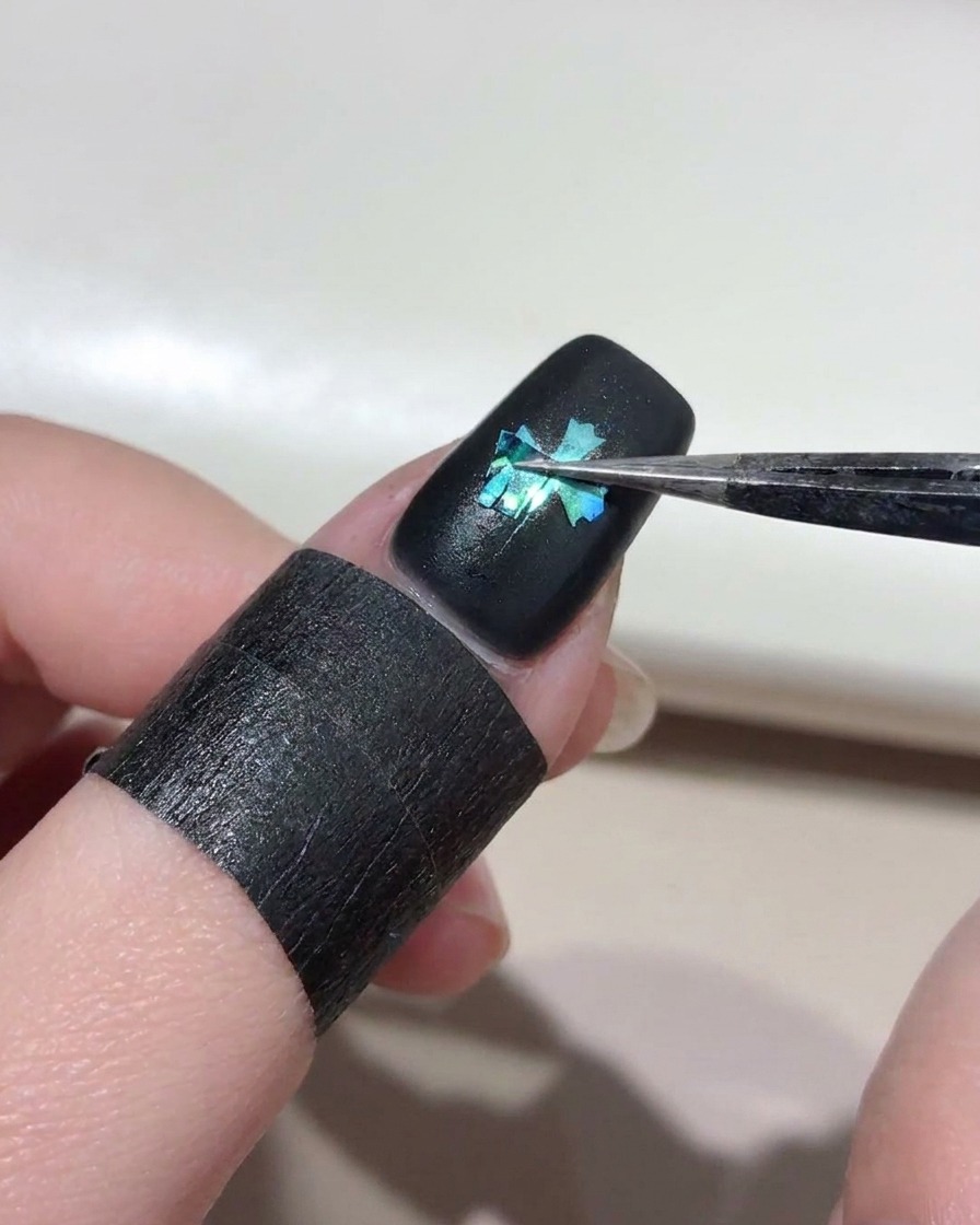 Eine Pinzette zieht vorsichtig eine Kreuzschablone von einem schwarz lackierten Nagel ab und enthüllt darunter eine hellblaue Cat-Eye-Kreuzform.