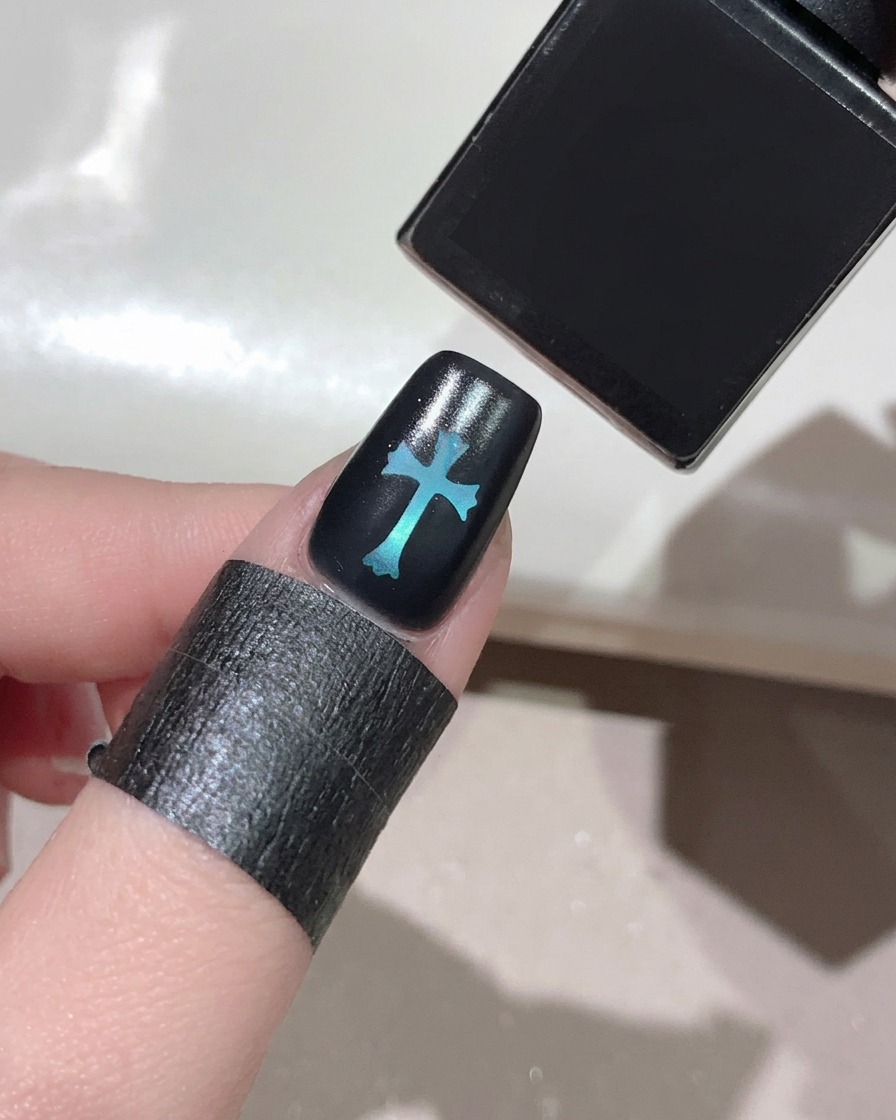 Ein Pinsel trägt einen klaren, glänzenden Top Coat über einem schwarzen Nagel mit einem leuchtenden blauen Kreuzdesign in der Mitte auf.