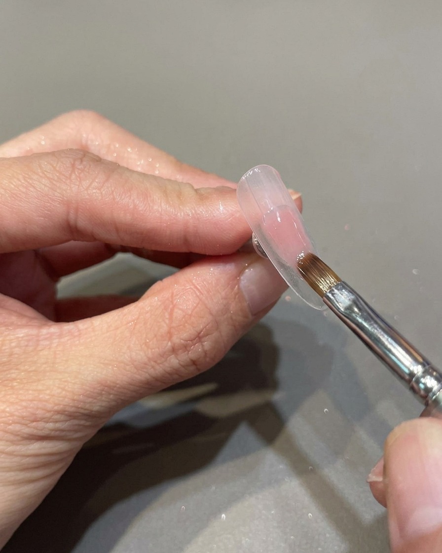 Ein professioneller Nagelpinsel trägt ein transparentes, rosa Gel auf einen vorbereiteten Nagel auf.