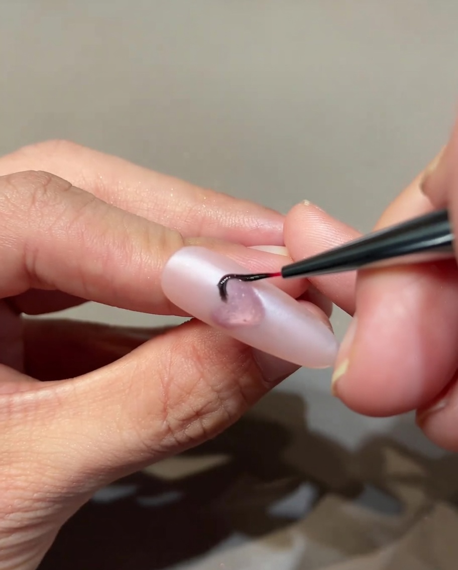 Ein dünner Nailart-Pinsel zeichnet feine schwarze Flammen auf einen rosa Nagel.
