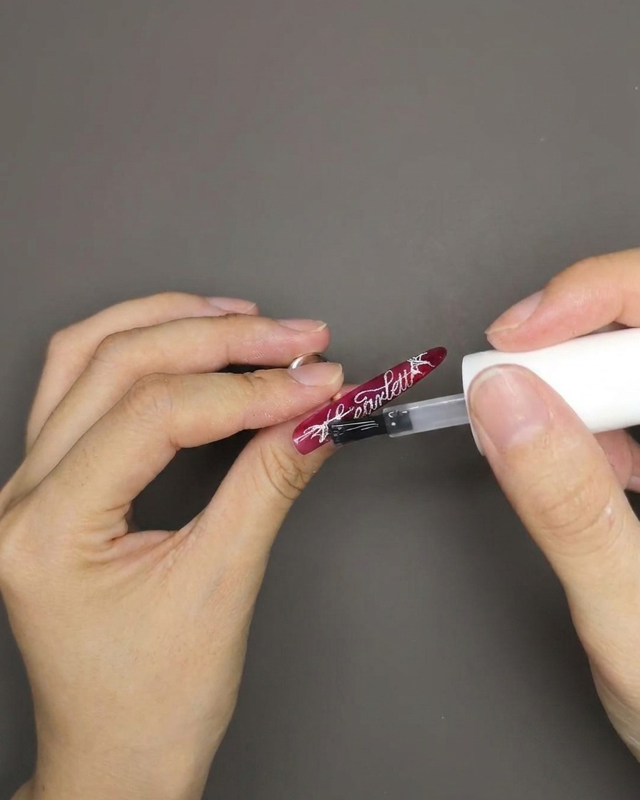 Ein Pinsel trägt einen glänzenden Top Coat auf einen Nagel mit rotem Cat-Eye-Effekt und weißem Schriftzug auf.