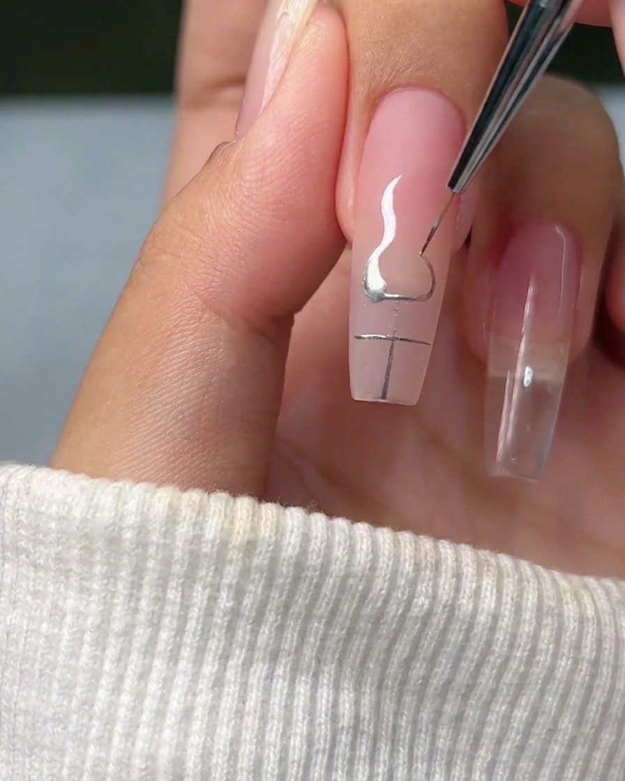 Ein Liner-Pinsel zeichnet elegante, flammenartige Kurven, die von den Enden eines silbernen Kreuzes auf einem Fingernagel ausgehen.