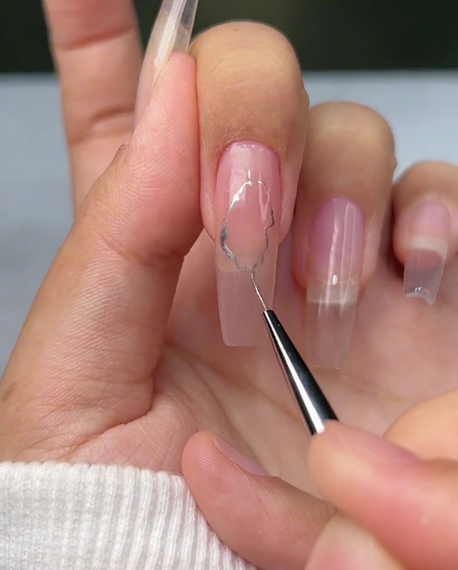 Ein feiner Liner-Pinsel zeichnet unregelmäßige, wellenförmige silberne Linien auf einen Nagel.