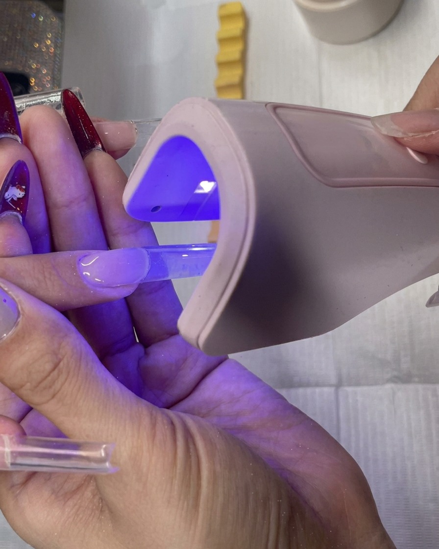 Ein Nageldesigner hält eine kleine LED-Handlampe über die klare quadratische Gel-Verlängerung zum Aushärten.