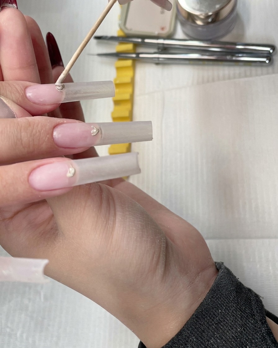 Ein Holzstäbchen platziert eine kleine Kugel Nude-Concealer-Gel am Nagelhautbereich einer Verlängerung.