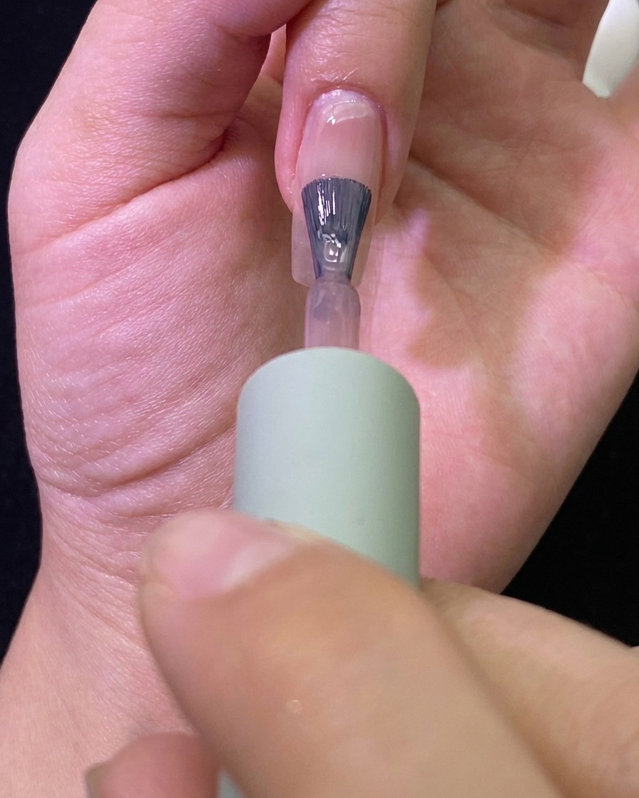 Nahaufnahme eines Pinsels, der ein transparentes Nude-Gel auf einen Naturnagel aufträgt.