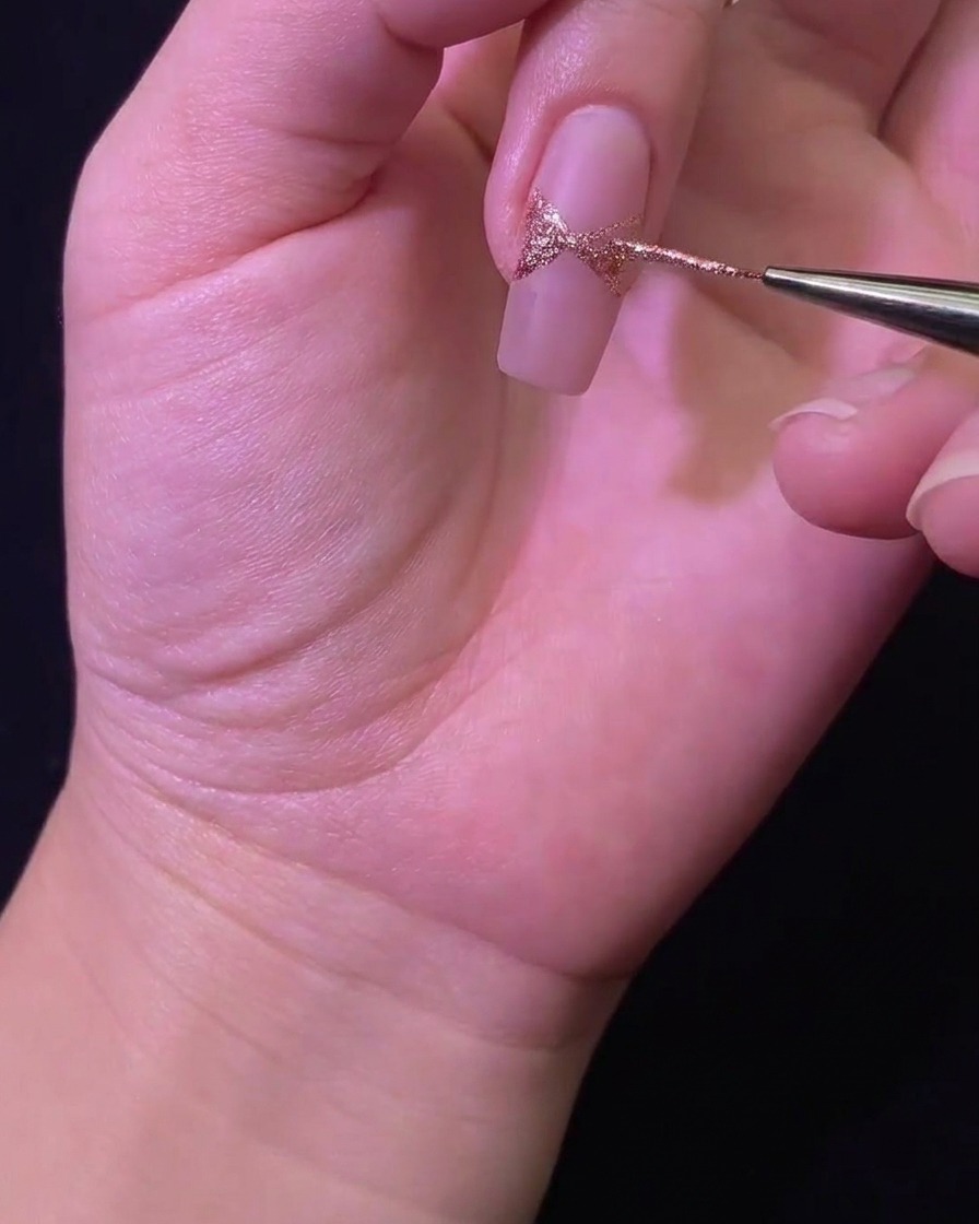 Nahaufnahme eines Liner-Pinsels, der die Glitzerschleifenform auf der Nageloberfläche ausfüllt.