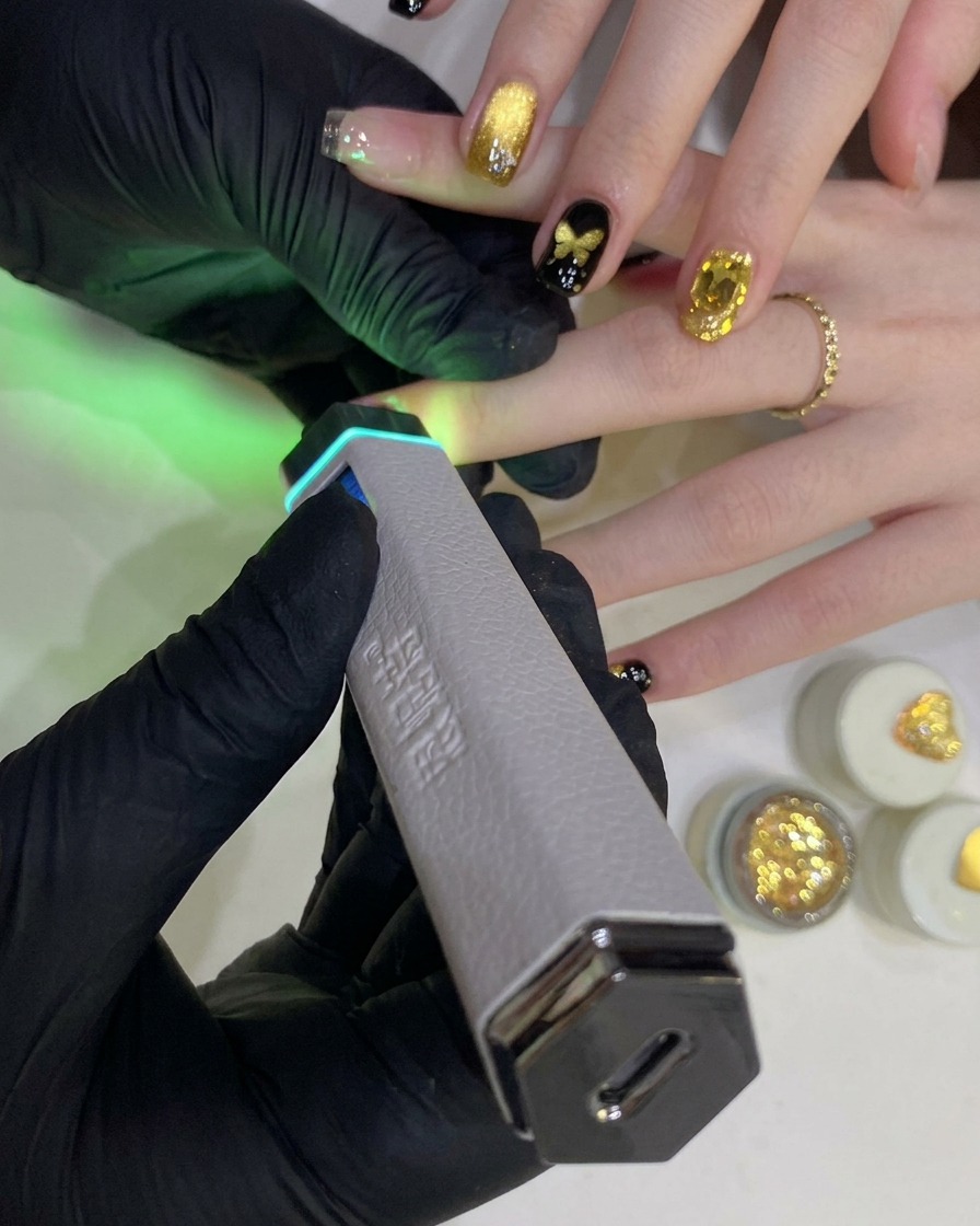 Eine behandschuhte Hand hält eine tragbare UV-/LED-Lampe über einen mit schwarzem Farbgel lackierten Fingernagel.