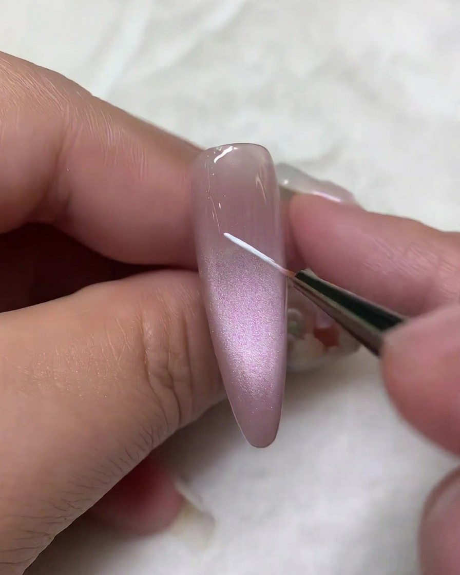 Nahaufnahme eines Fine Liner Pinsels, der eine dünne weiße vertikale Linie in die Mitte eines malvenfarbenen Cat-Eye-Nagels zieht.