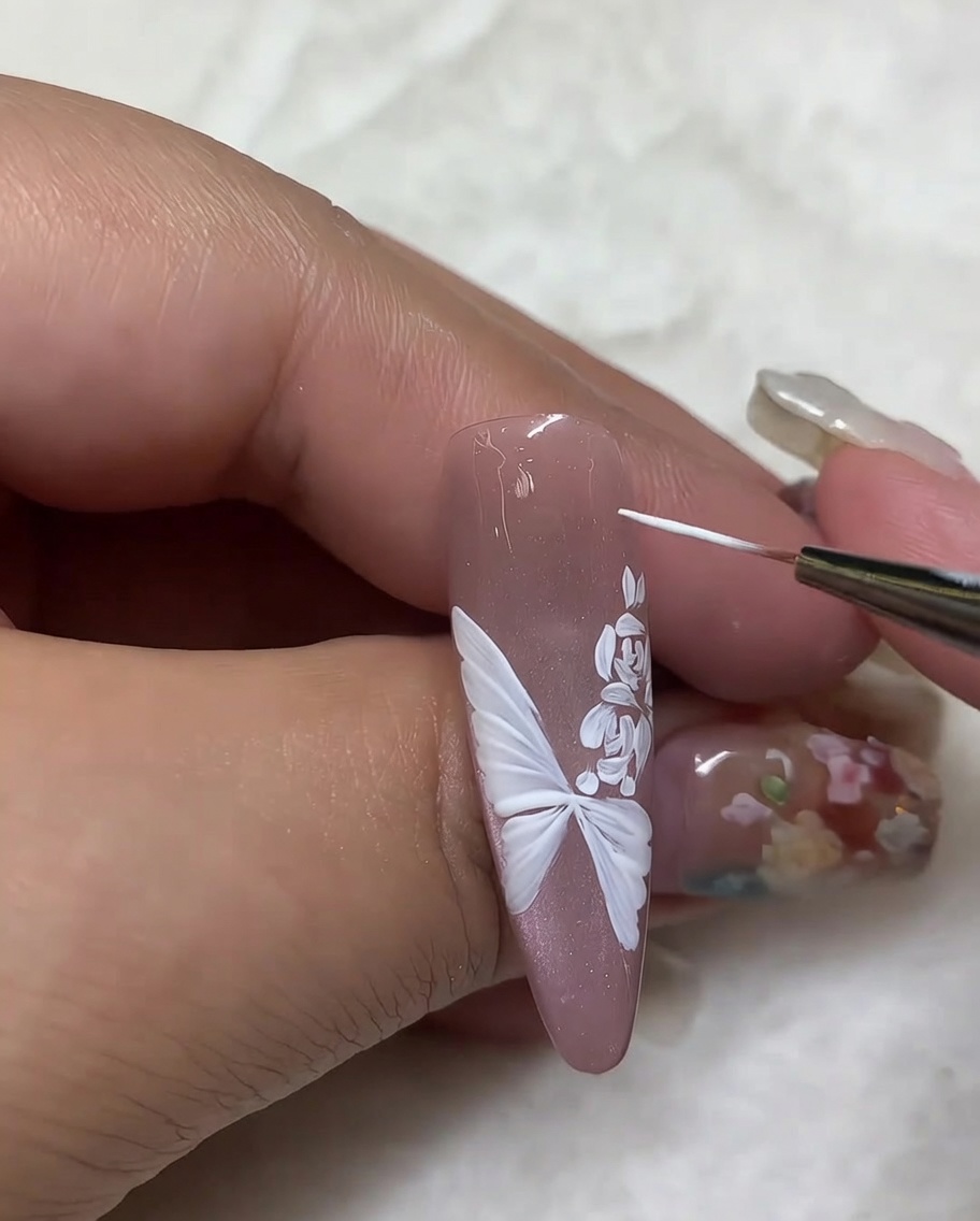 Ein feiner Pinsel fügt kleine weiße Highlight-Punkte rund um das Schmetterlings- und Rosendesign auf einem nudefarbenen Nagel hinzu.