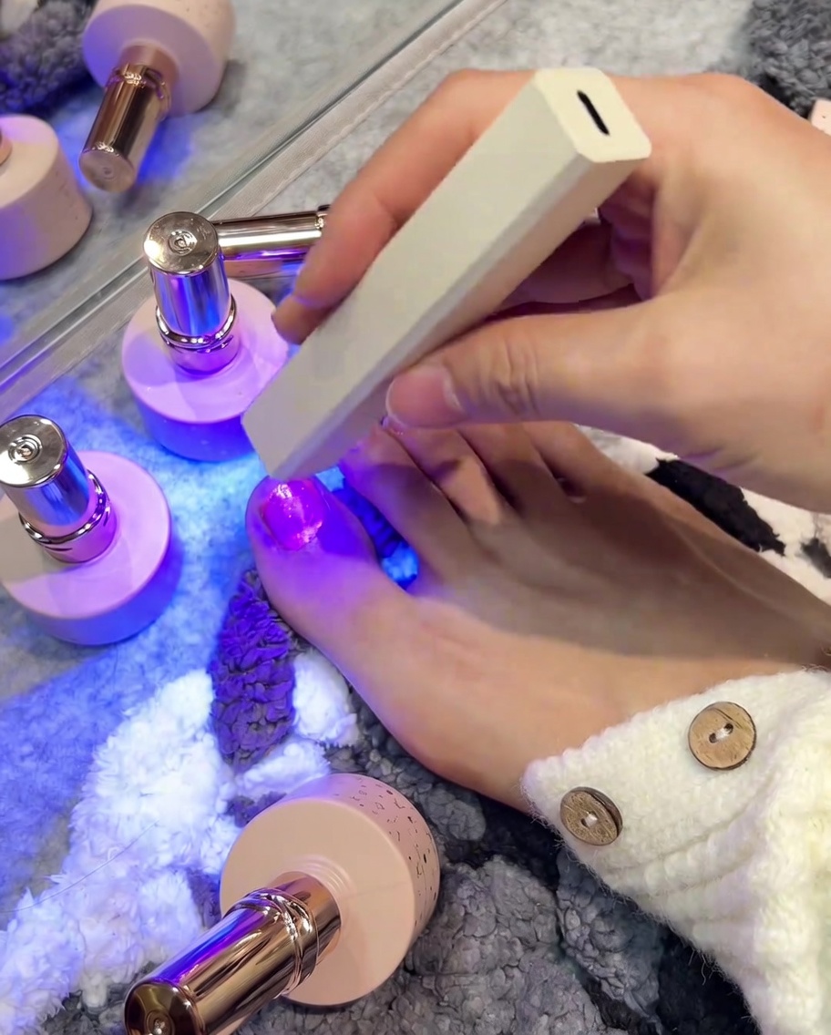 Eine Hand hält eine kleine weiße, rechteckige UV/LED-Lampe direkt über einen frisch lackierten rechten großen Zehennagel, um das Gel auszuhärten.