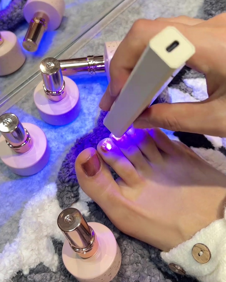 Eine handgehaltene UV/LED-Lampe ist über einem rosafarbenen Zehennagel positioniert, wobei das violette Licht den Nagel während des Aushärtens beleuchtet.
