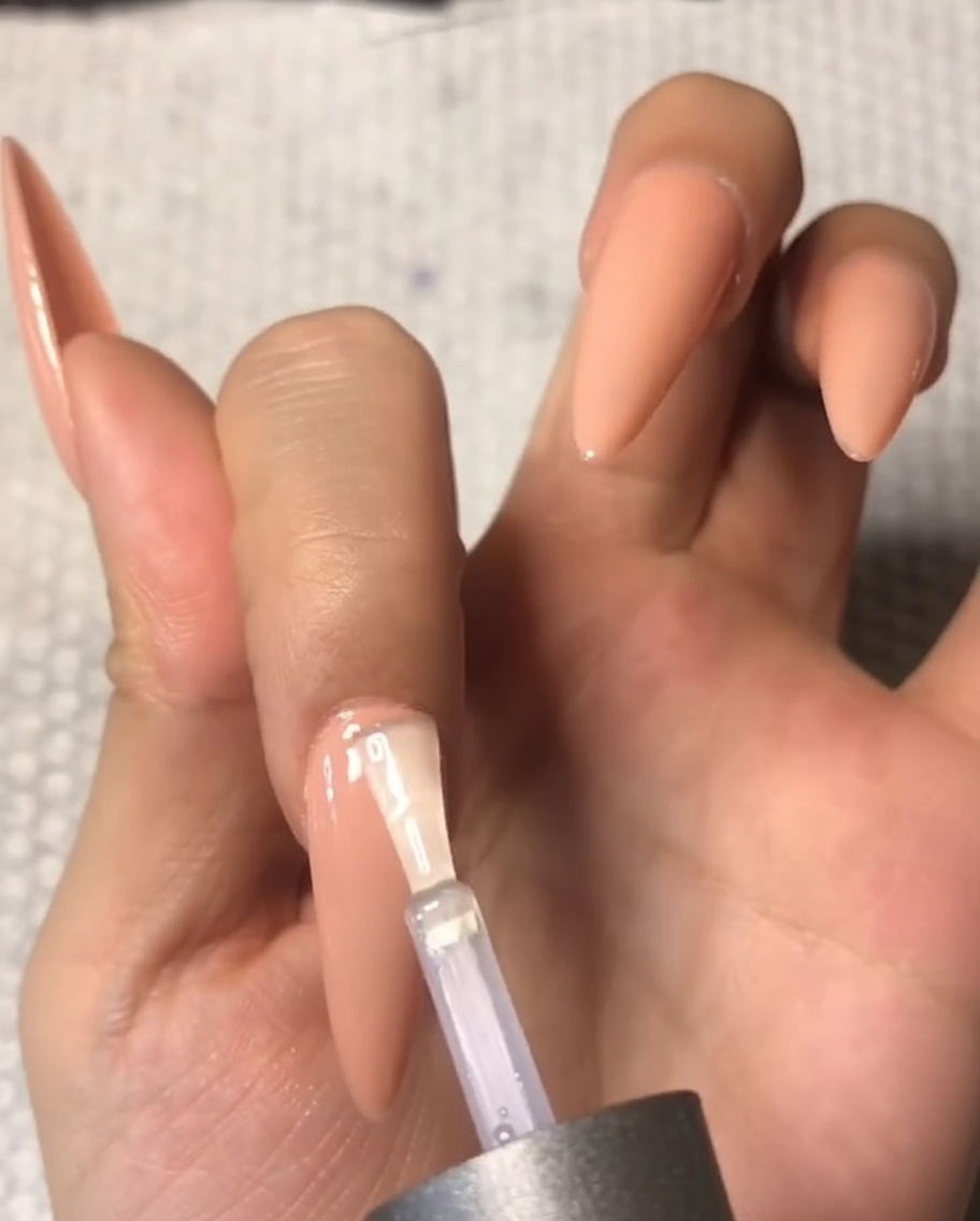 Nahaufnahme eines Pinsels, der vorsichtig klaren, gehärteten Top Coat nahe der Nagelhaut eines mandelförmigen Nude-Nigels aufträgt.