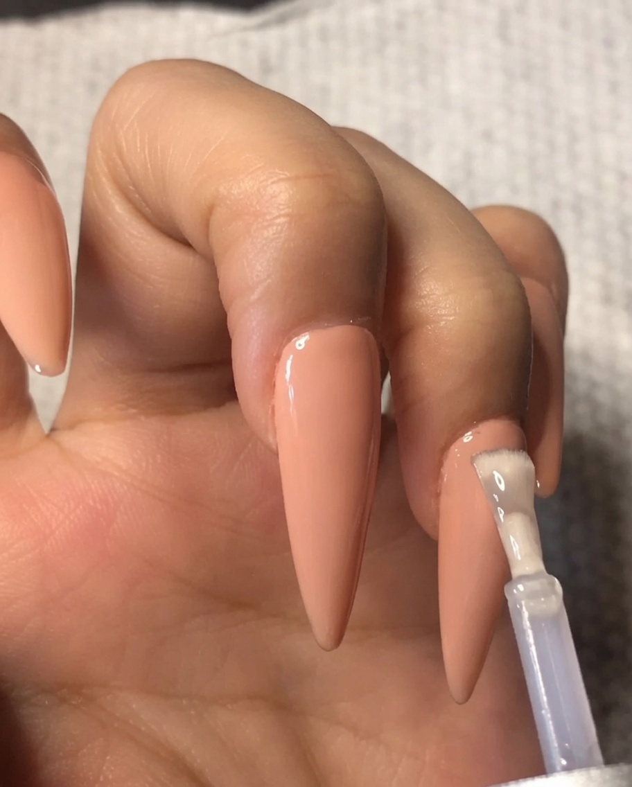 Ein Pinsel trägt eine Schicht klares Gel-Top-Coat über den Nude-Nagellack auf dem Ringfinger auf.