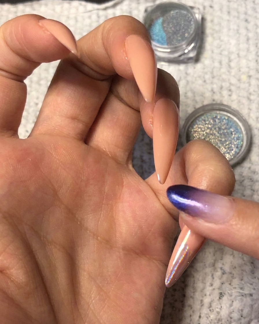 Ein Finger drückt leuchtend blau-lila holografisches Chrom-Puder auf die Spitze eines mandelförmigen Nude-Nigels.