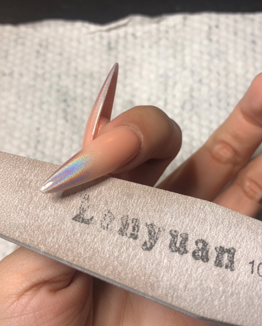 Eine Hand benutzt eine feine Schwammfeile, um sanft den freien Rand eines Stiletto-Nagels mit holografischem Chrom-Puder zu glätten.