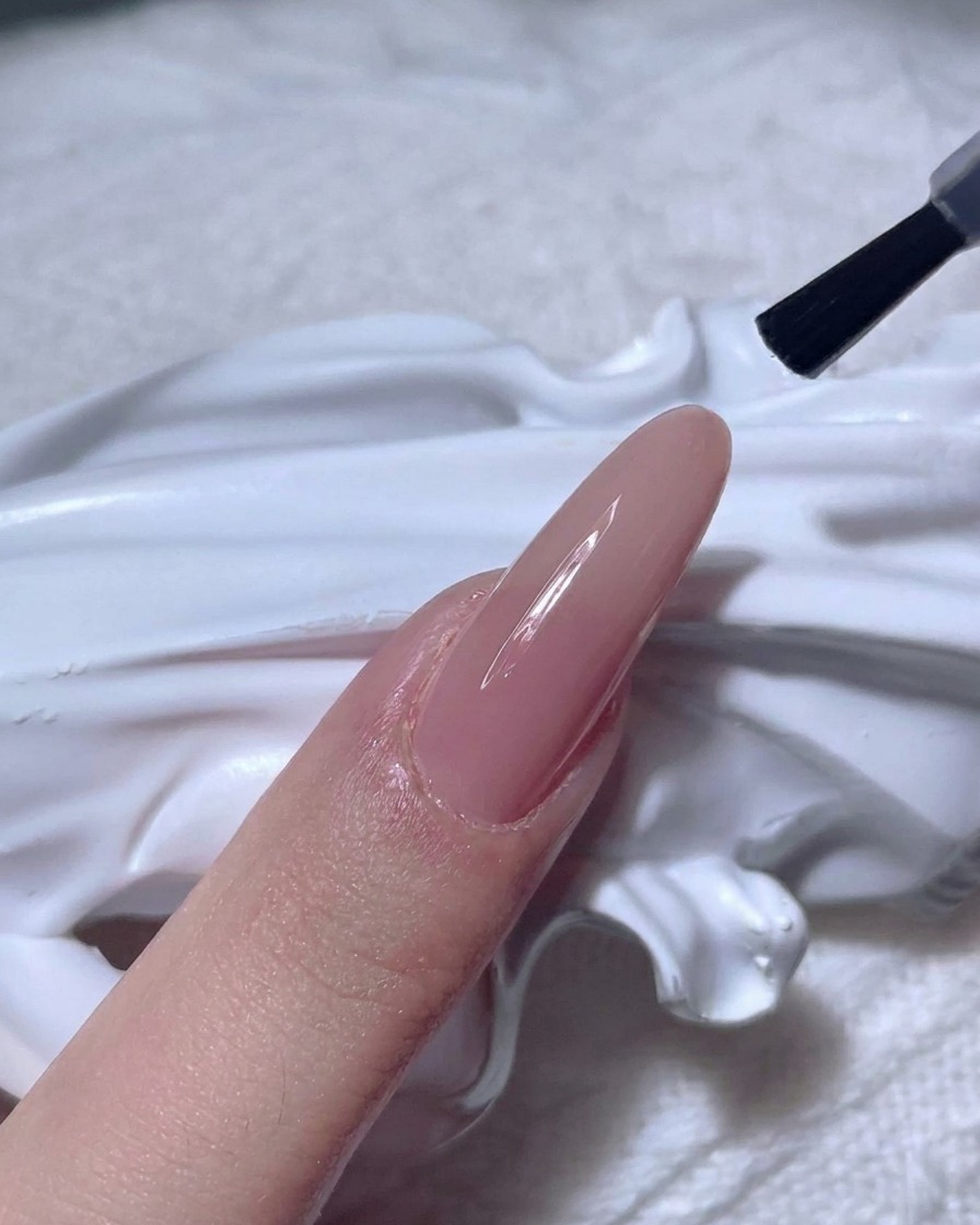 Ein Nagelpinsel trägt einen klaren, glänzenden Top Coat auf einen Nude-farbenen Nagel auf.