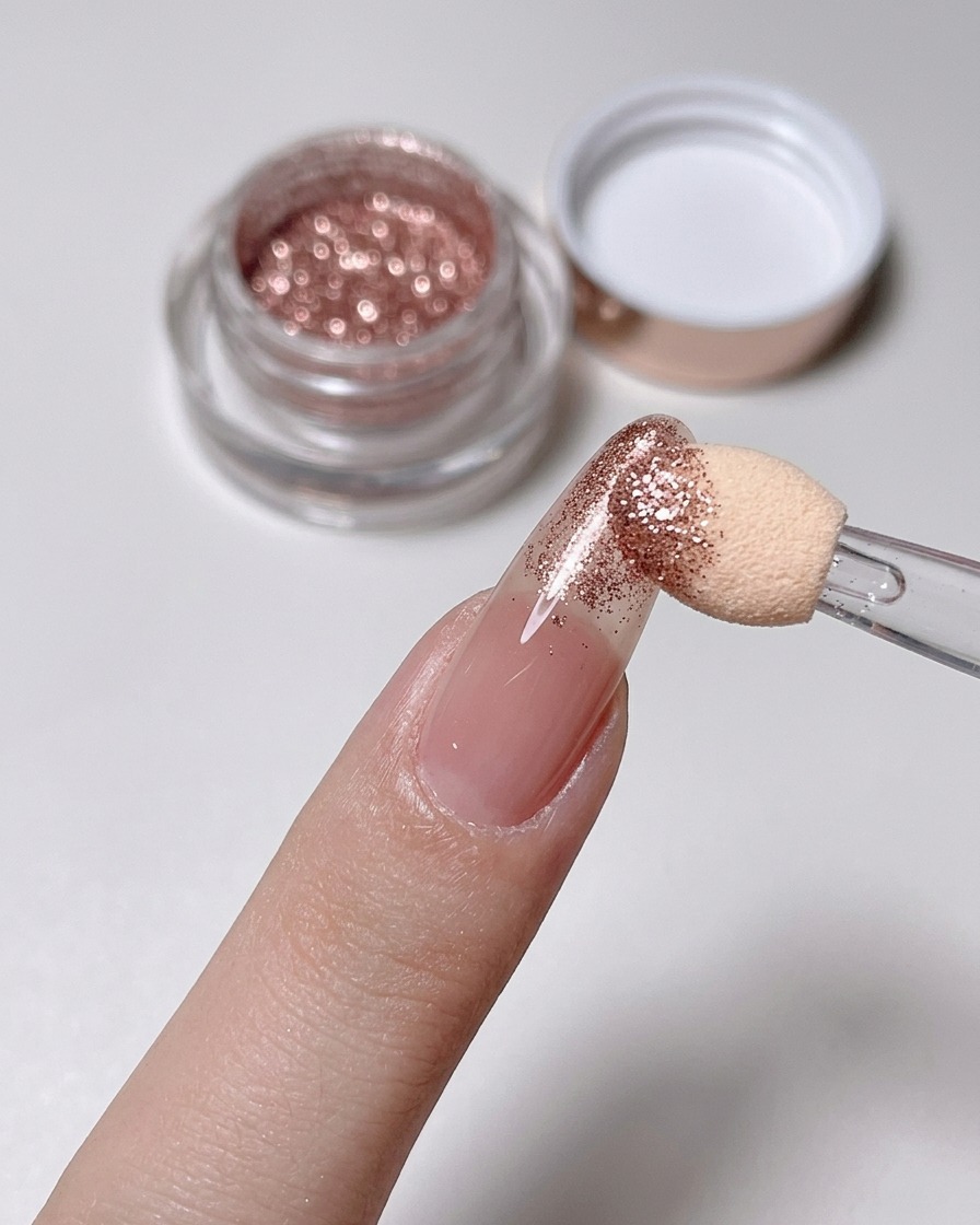 Ein Schwamm-Applikator poliert Roségold-Puder auf die Spitze eines Nude-Nagels.
