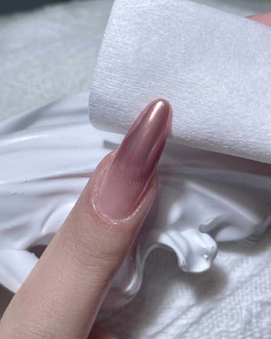 Eine Hand nutzt ein fusselfreies Pad, um die Ränder eines Roségold-Chrome-Nagels zu säubern.