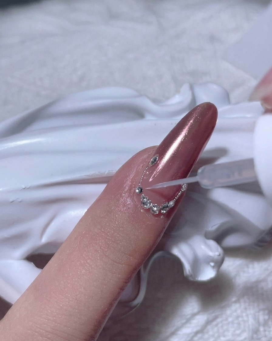 Ein vollendeter Bogen aus kleinen klaren Strasssteinen entlang der Nagelhautlinie eines Roségold-Chrome-Nagels.