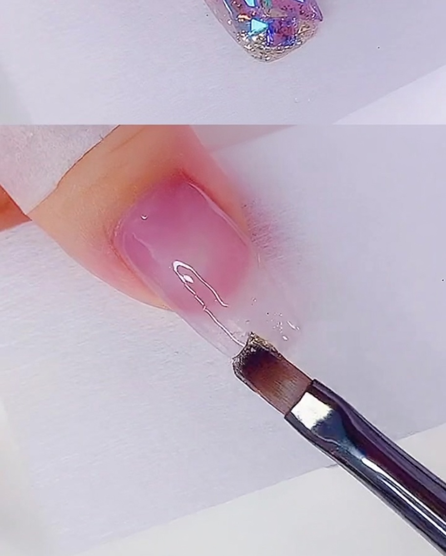 Ein Flachpinsel trägt eine dünne Schicht roségoldenes Glitzergel auf die Nagelspitze auf.