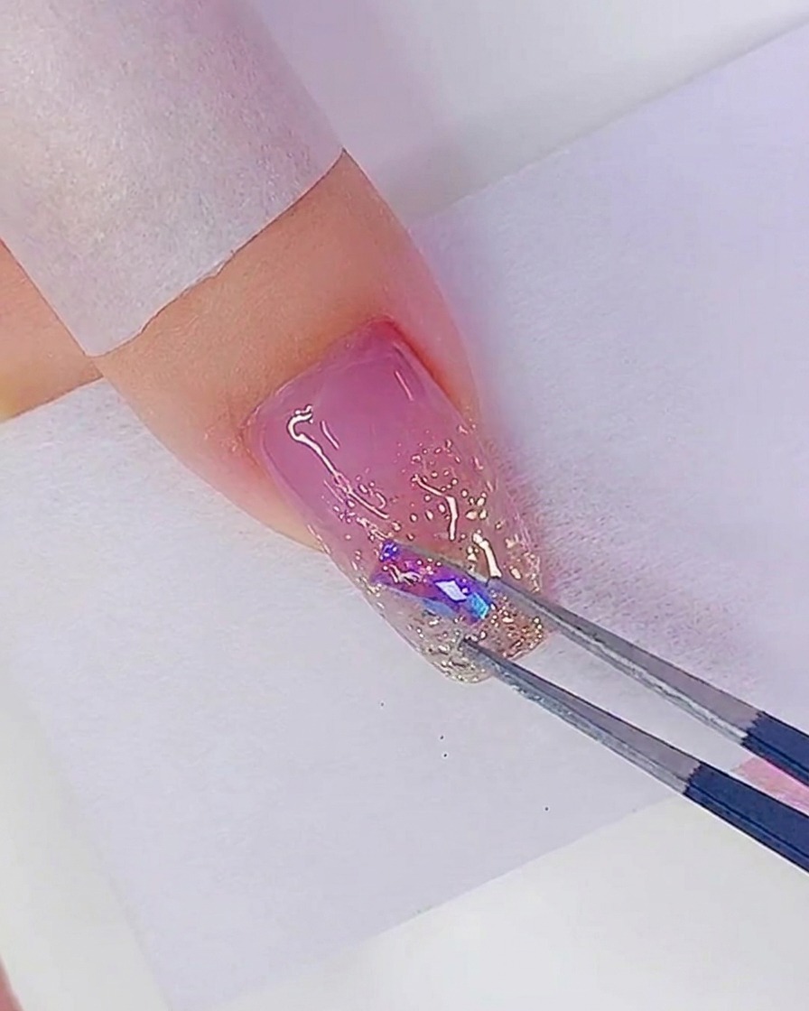 Eine Pinzette setzt einen irisierenden Strassstein auf einen Klebepunkt auf dem glitzernden Nagel.