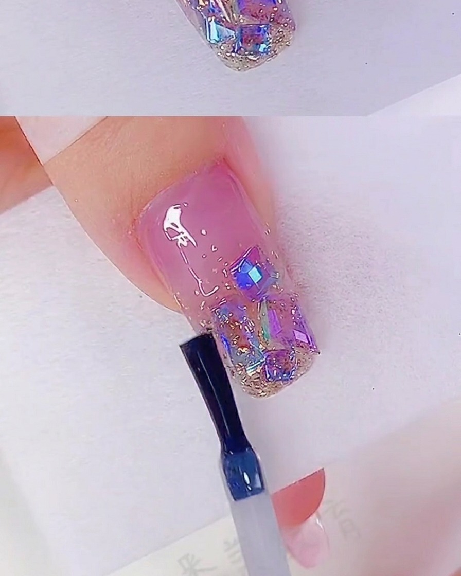 Ein Nagelpinsel trägt einen klaren Top Coat über den Nagel auf, der mit Glitzer und Strasssteinen verziert ist.