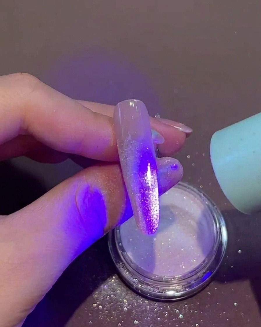Eine Hand hält eine kleine tragbare UV/LED-Lampe direkt über einen Fingernagel, der feuchtes Gel und loses Puder an der Spitze aufweist.