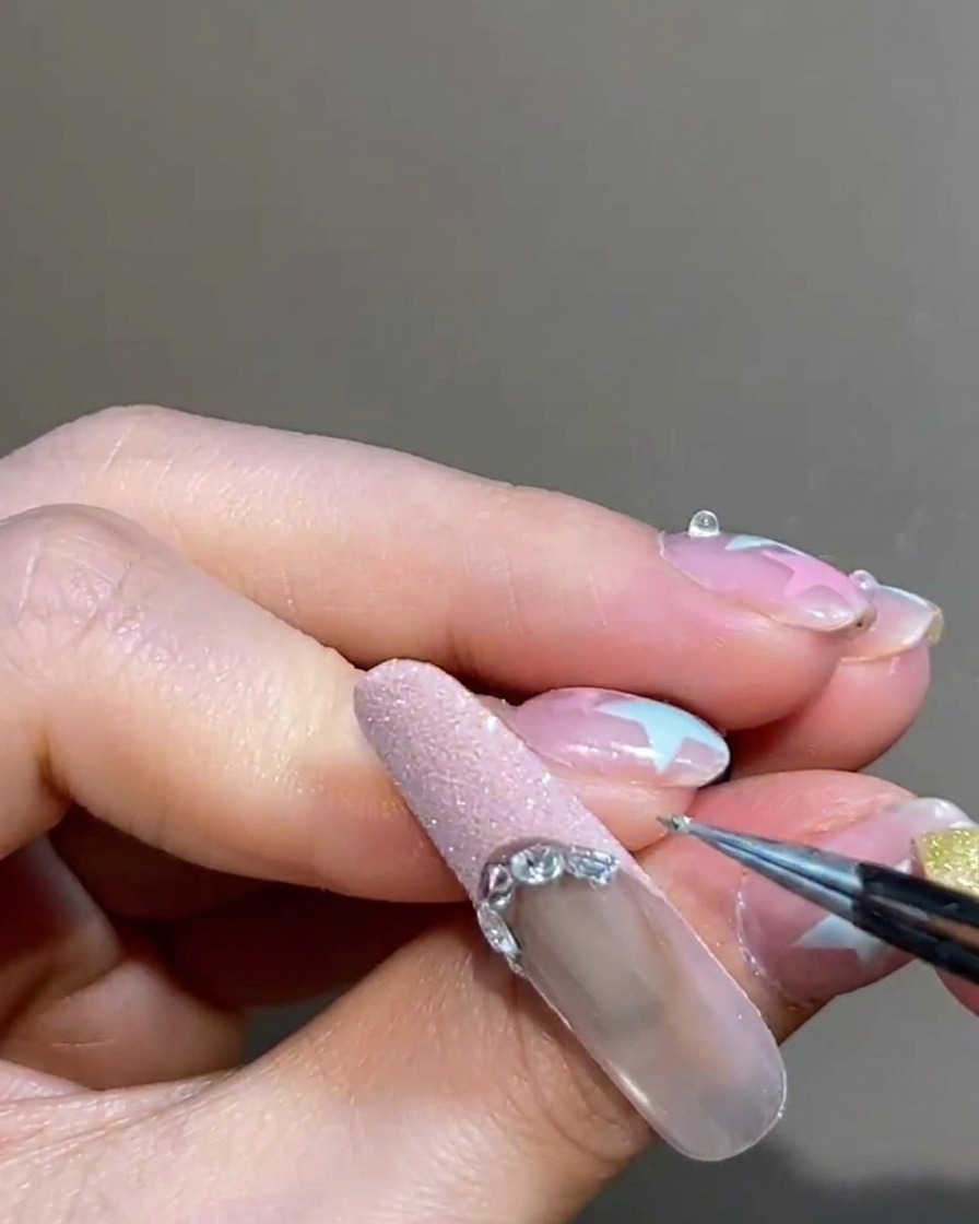 Eine Nahaufnahme einer Pinzette, die ein kleines silbernes Metallkügelchen entlang der Begrenzung einer rosa French-Spitze platziert.