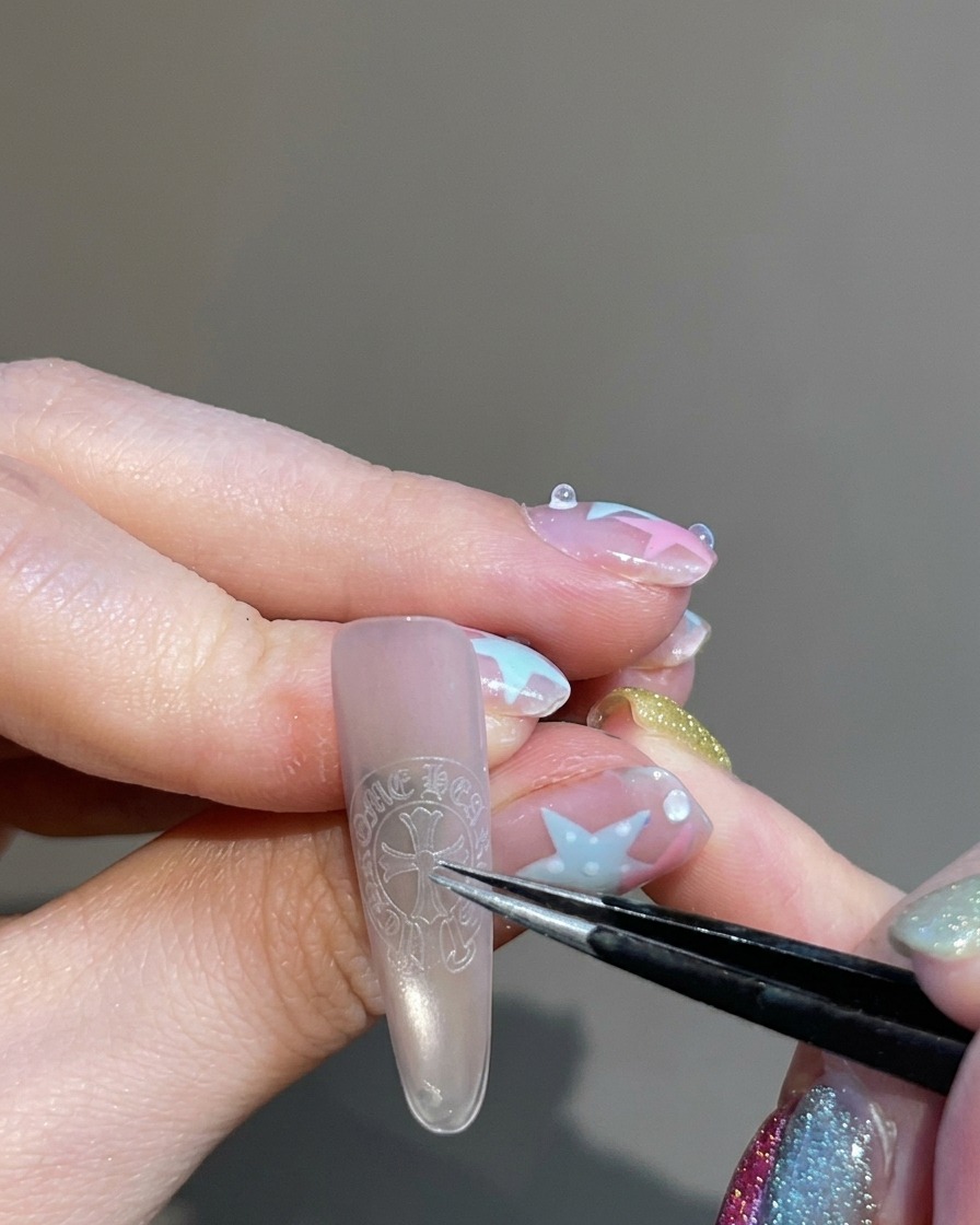 Eine Pinzette hält und platziert einen kreuzförmigen Sticker auf die Mitte eines Fingernagels.
