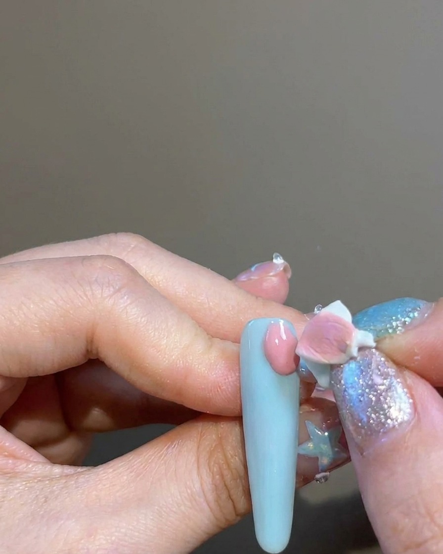 Ein Finger benutzt einen kleinen Schwamm-Applikator, um rosa Pigment auf den Nagel um einen kreuzförmigen Sticker zu tupfen.